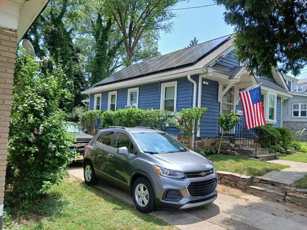 A gray SUV parked in a driveway in front of a blue house with solar panels and an American flag.
