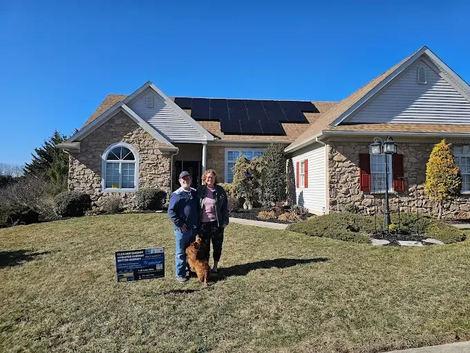 Couple and dog stand in front of a house with solar panels on the roof and a sign in the yard on a sunny day.