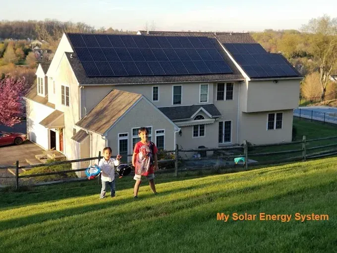 Two children run on a grassy hill in front of a house with solar panels.