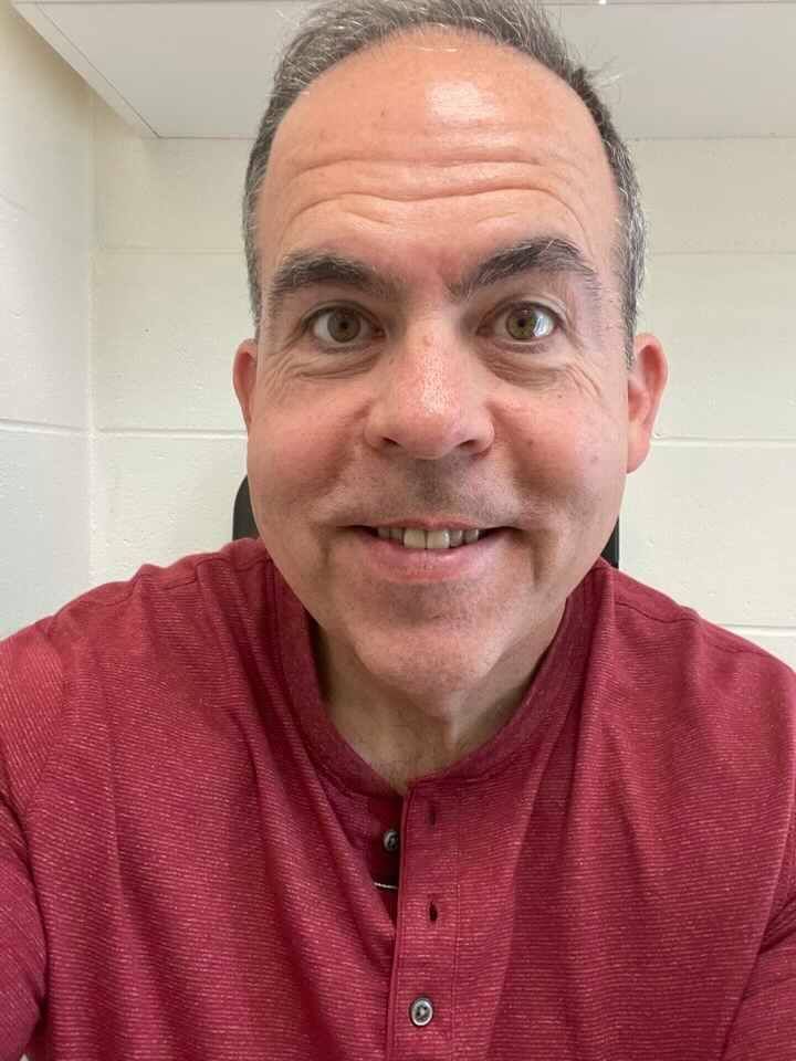 Man in red shirt smiling, close-up. Light-colored background.