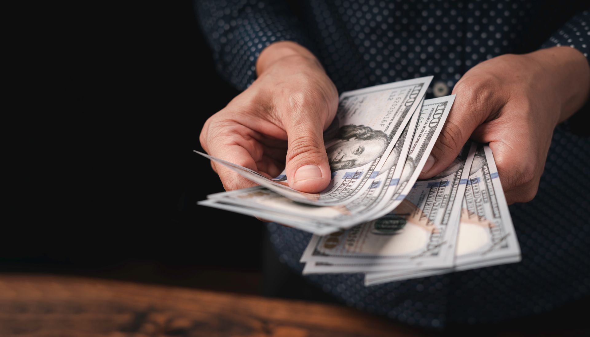Person holding and counting several hundred-dollar bills on a wooden table.