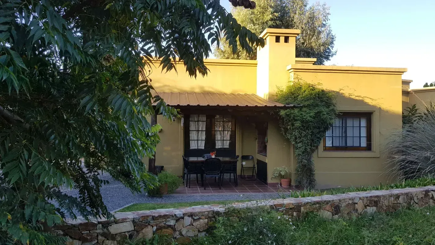 Casa amarilla con porche sombreado, chimenea y sillas de patio detrás de árboles y vegetación.
