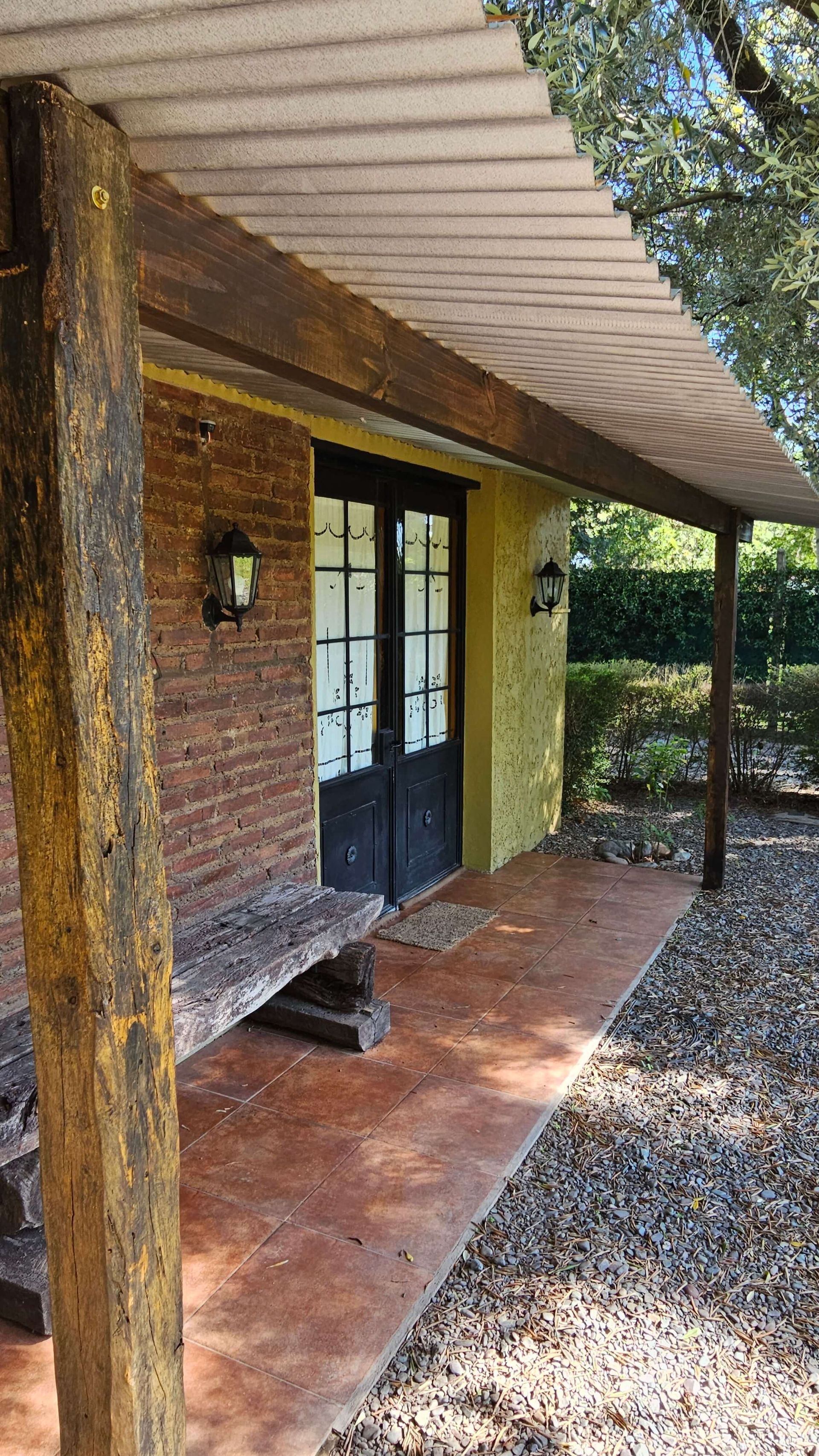 Entrada rústica de ladrillo con puertas dobles negras, paredes amarillas y un porche con baldosas bajo un alero.