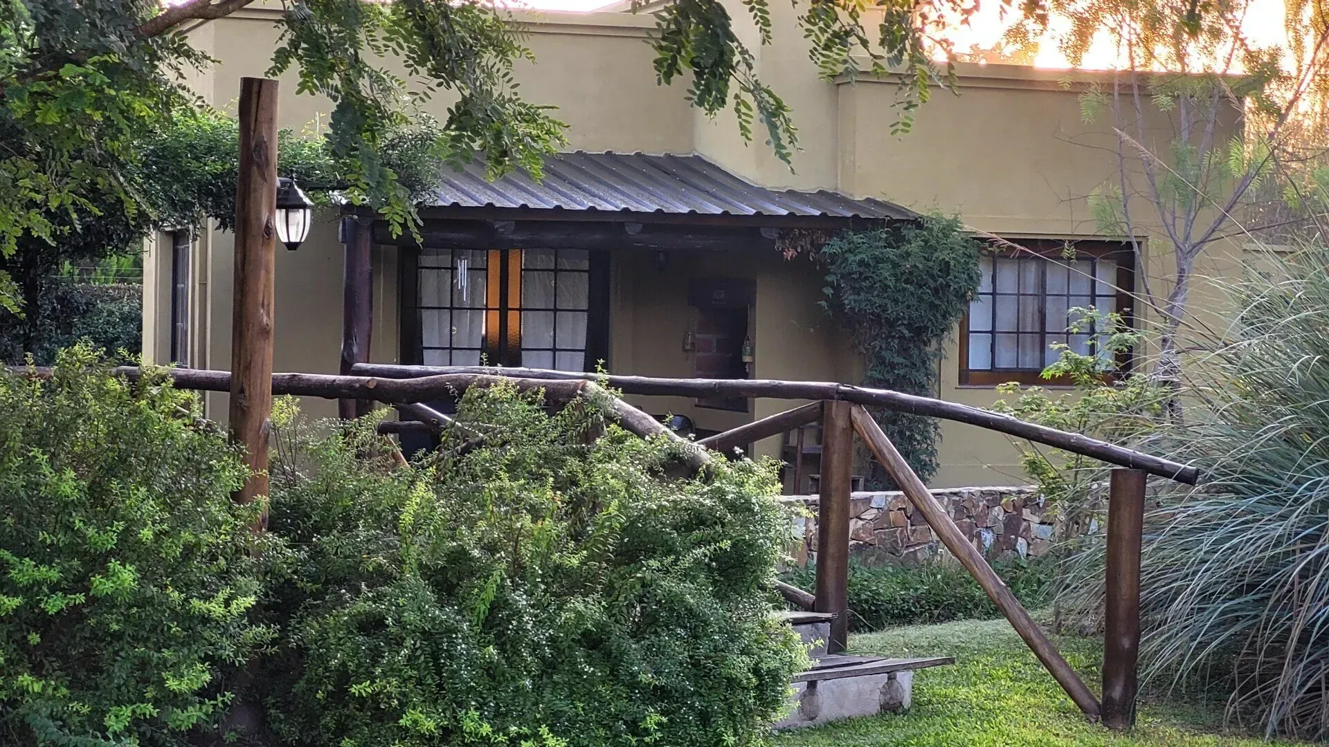 Casa rústica con porche, barandilla de madera y frondosos arbustos verdes en un jardín.