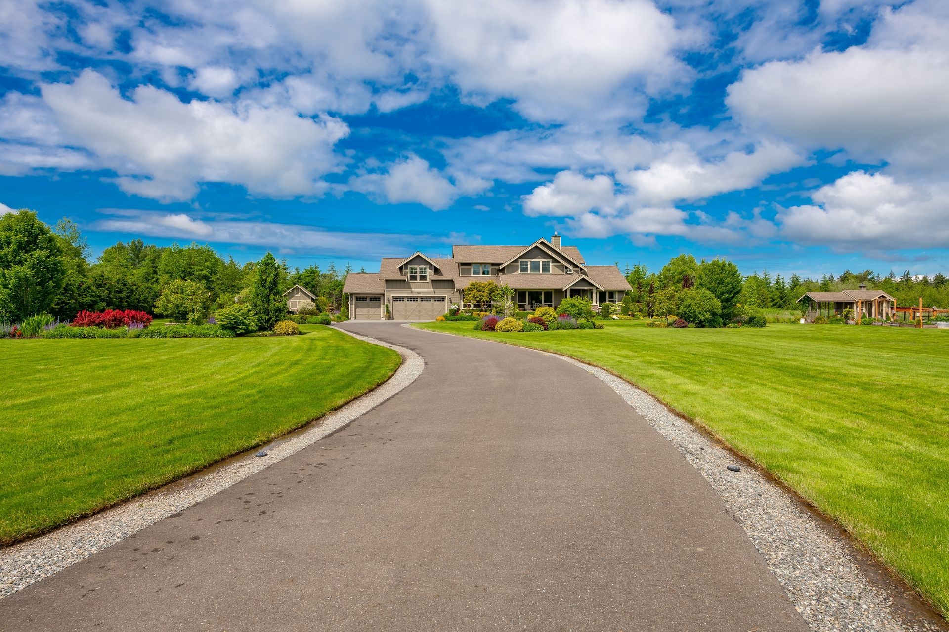 A paved driveway leads to a suburban house surrounded by green lawns under a bright blue sky with fluffy white clouds.