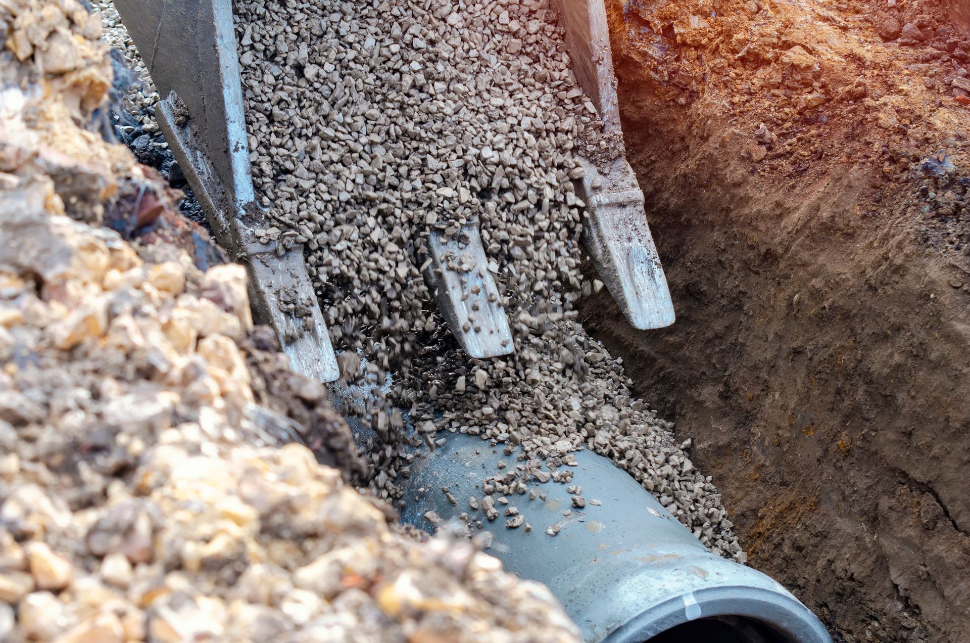 An excavator bucket pours gravel over a grey drainage pipe installed in an open dirt trench.