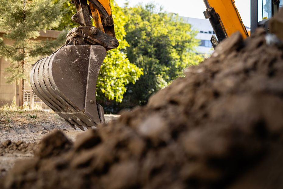 Yellow excavator bucket raised over a mound of dirt with trees in the background.
