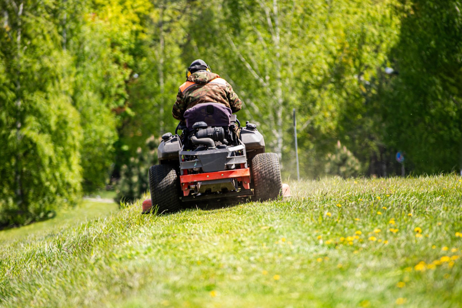 A person wearing a camouflage jacket operates a riding lawn mower across a sunny, grassy field lined with trees.