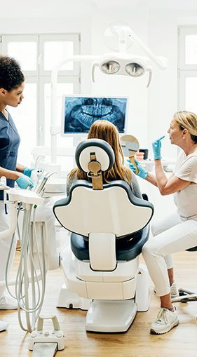 A woman is sitting in a dental chair while two female dentists work on her teeth.