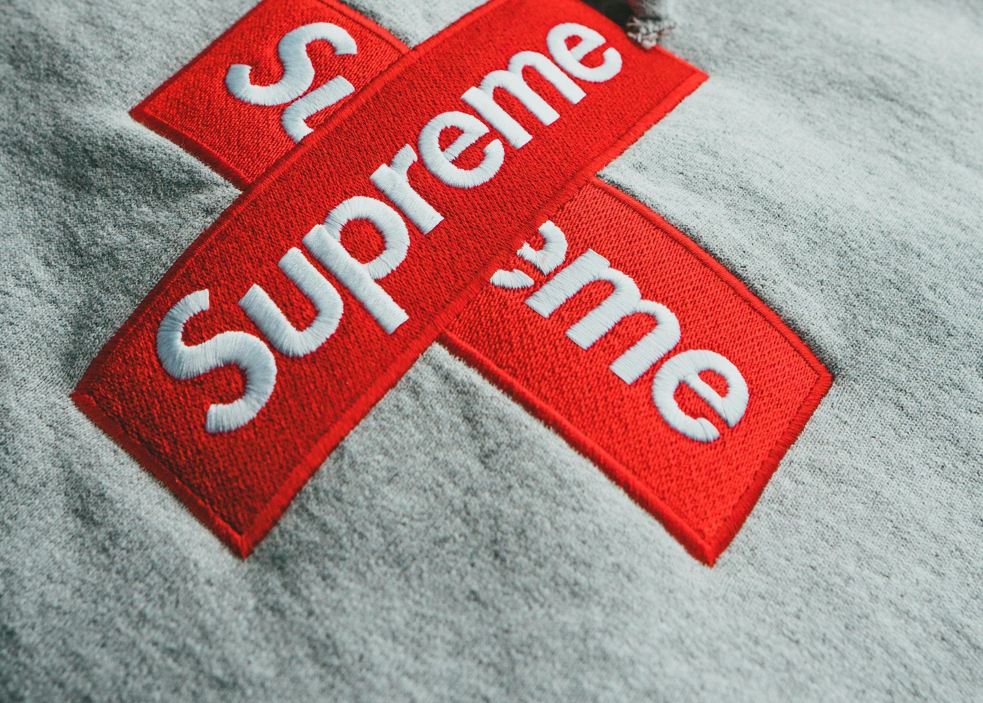 Embroidered red Supreme logo in a cross shape on a gray hoodie.
