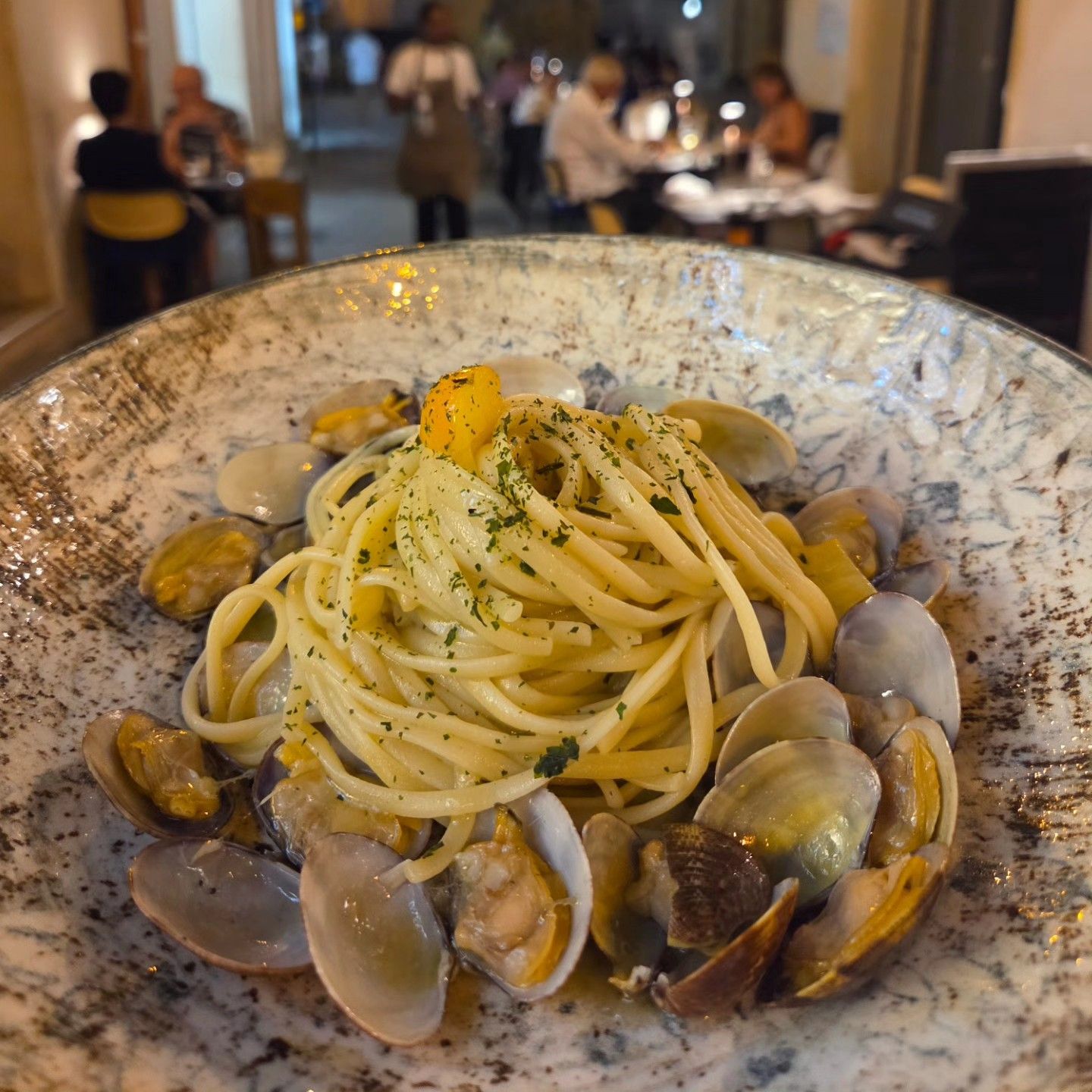 Un piatto di pasta alle vongole in una ciotola bianca, guarnito con erbe aromatiche, su un tavolo di ristorante.