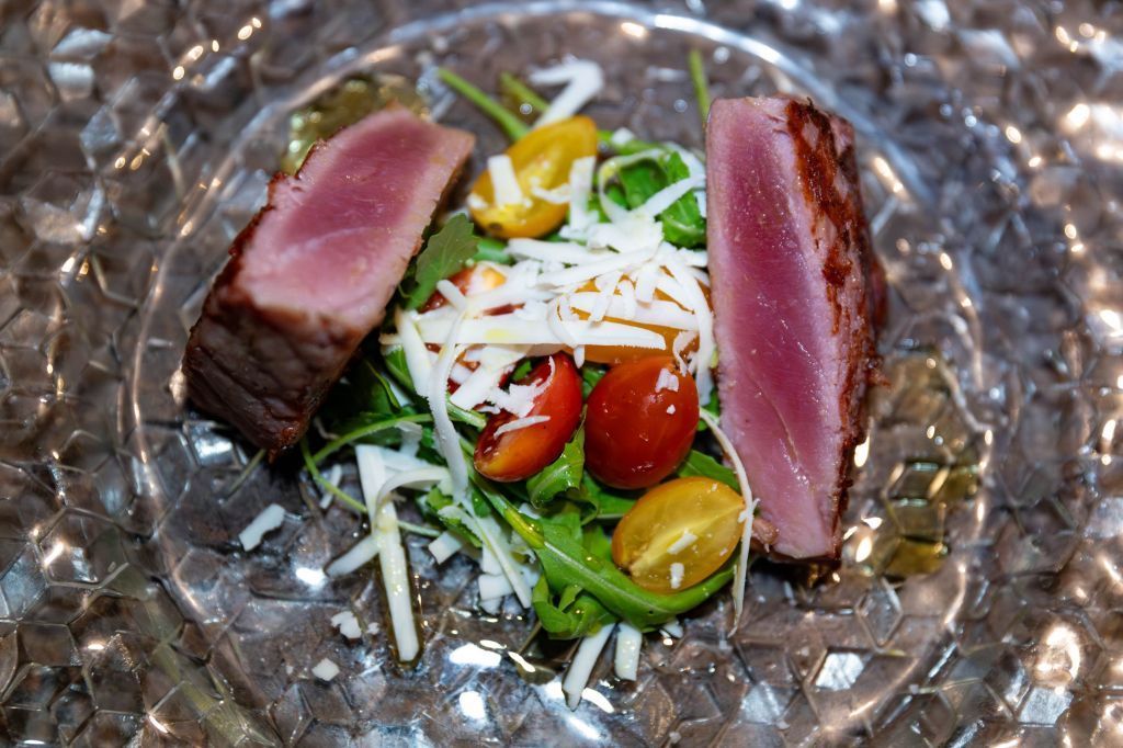 Fette di tonno scottato servite su un piatto di vetro, adagiate su un'insalata con pomodorini, verdure a foglia verde e formaggio grattugiato.