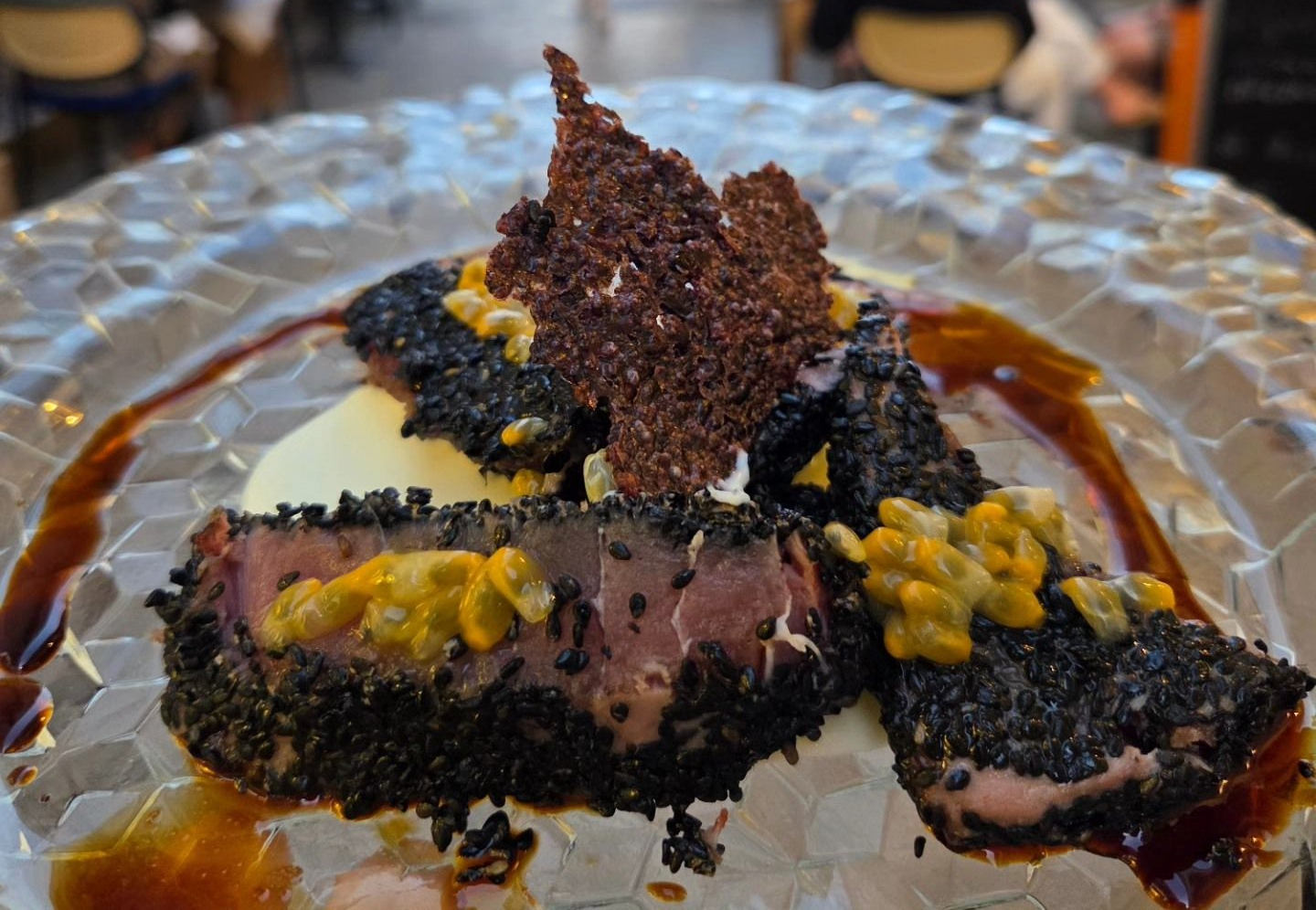 Dessert impiattato con copertura di briciole scure, salsa gialla e guarnizione di cioccolato su un piatto di vetro.