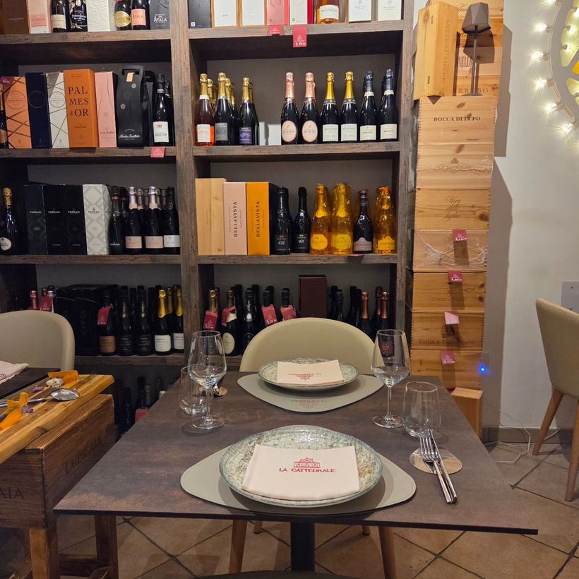 Tavolo da pranzo in un'enoteca, apparecchiato davanti a scaffali di bottiglie di vino, con piatti, bicchieri e lucine.