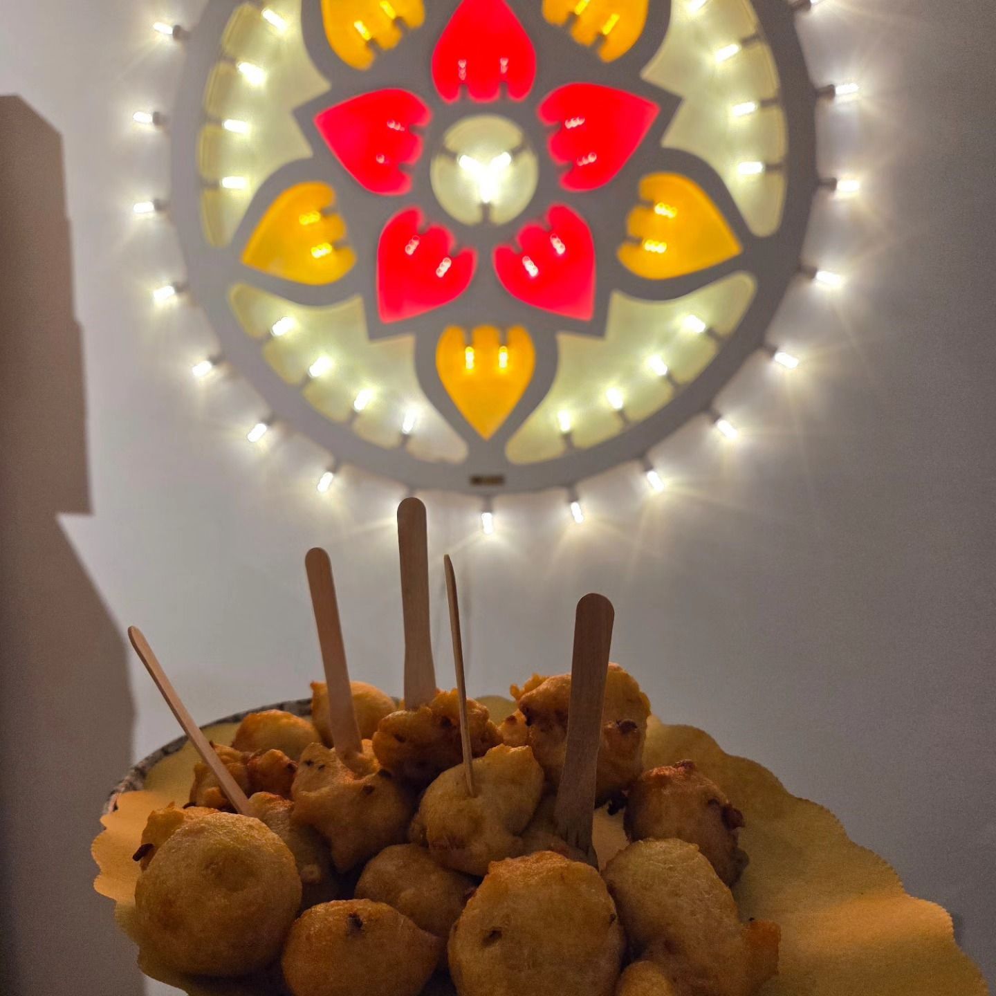 Piatto di snack fritti con spiedini di legno davanti a una luminosa lampada a parete a forma di fiori.