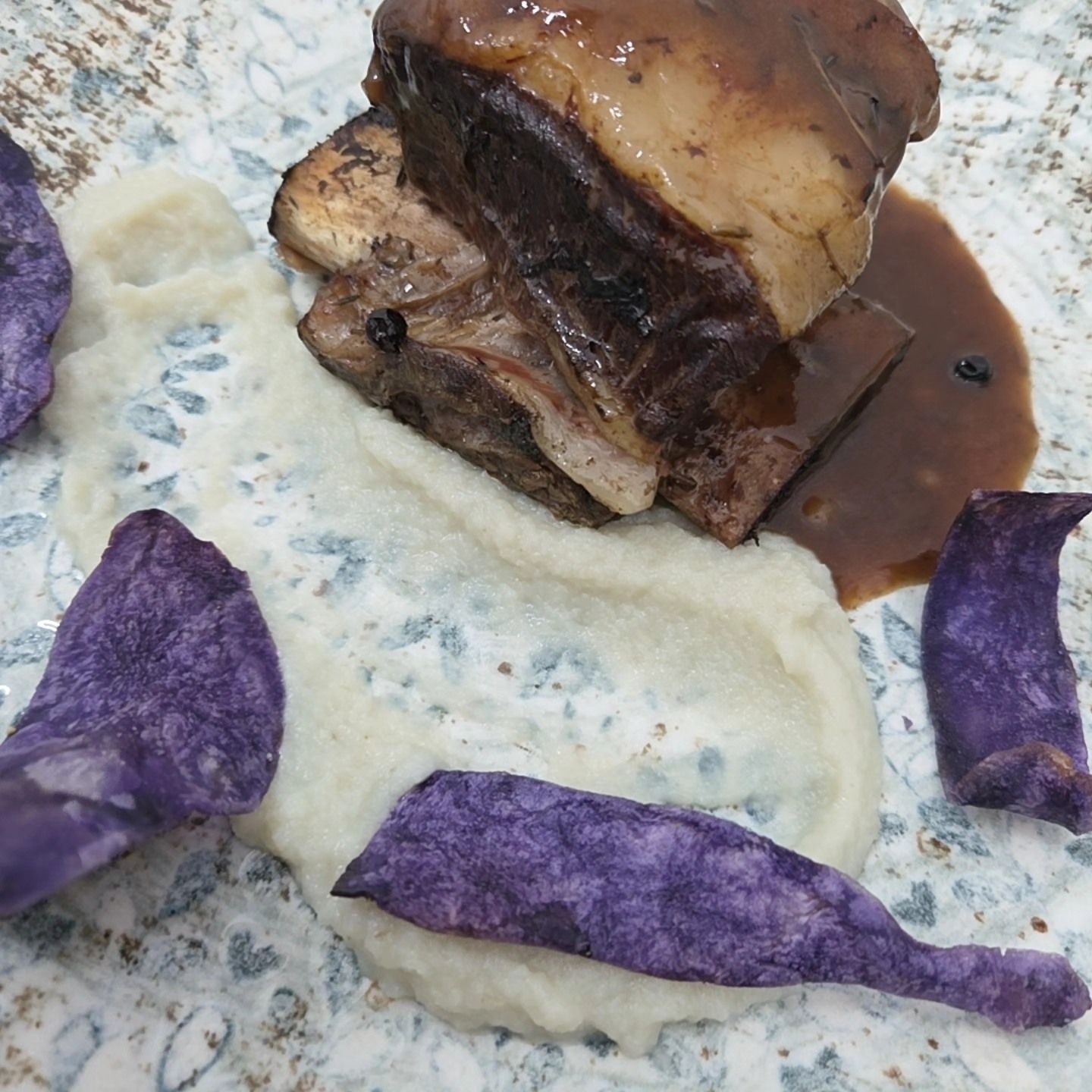 Carne brasata servita con salsa bruna su purè bianco, guarnita con chips di verdure viola su una superficie punteggiata.