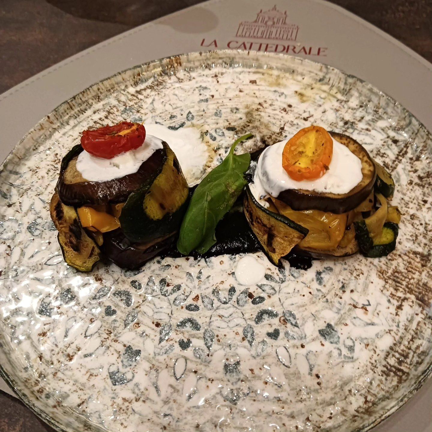 Due antipasti gourmet impilati su un piatto decorativo con crema bianca e guarnizione rossa, con verdure al centro.