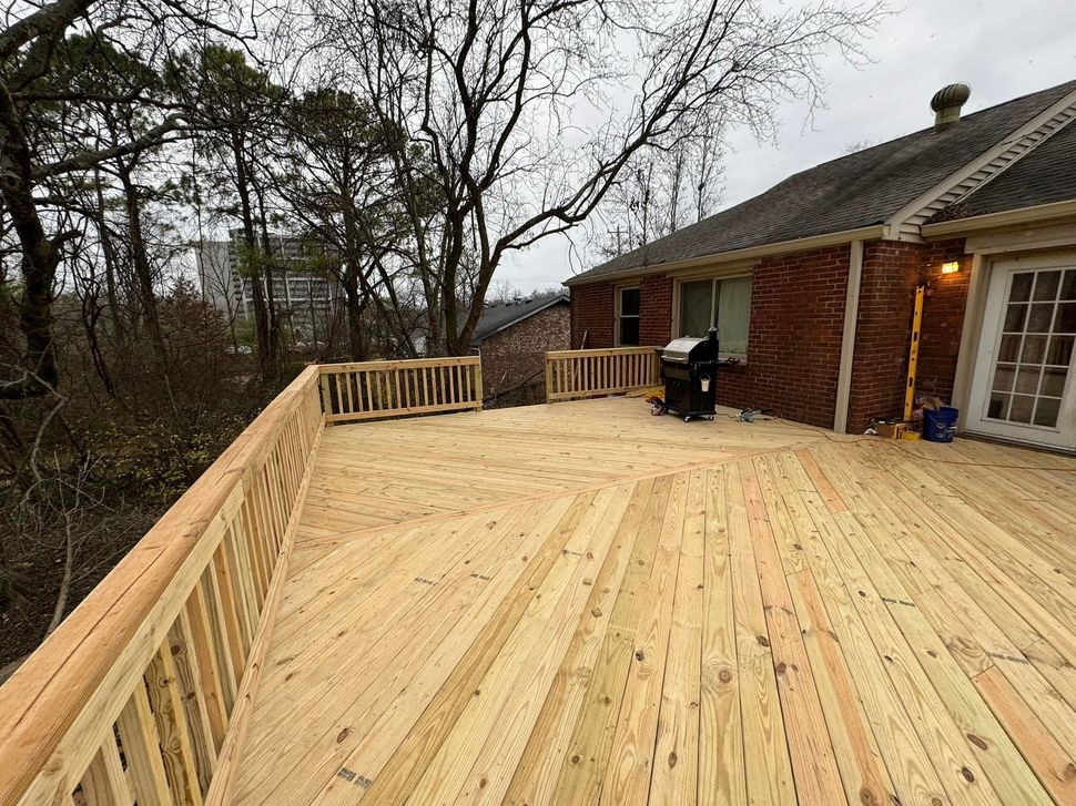 A newly built wooden deck with railings attached to a brick house, overlooking a wooded area on an overcast day.