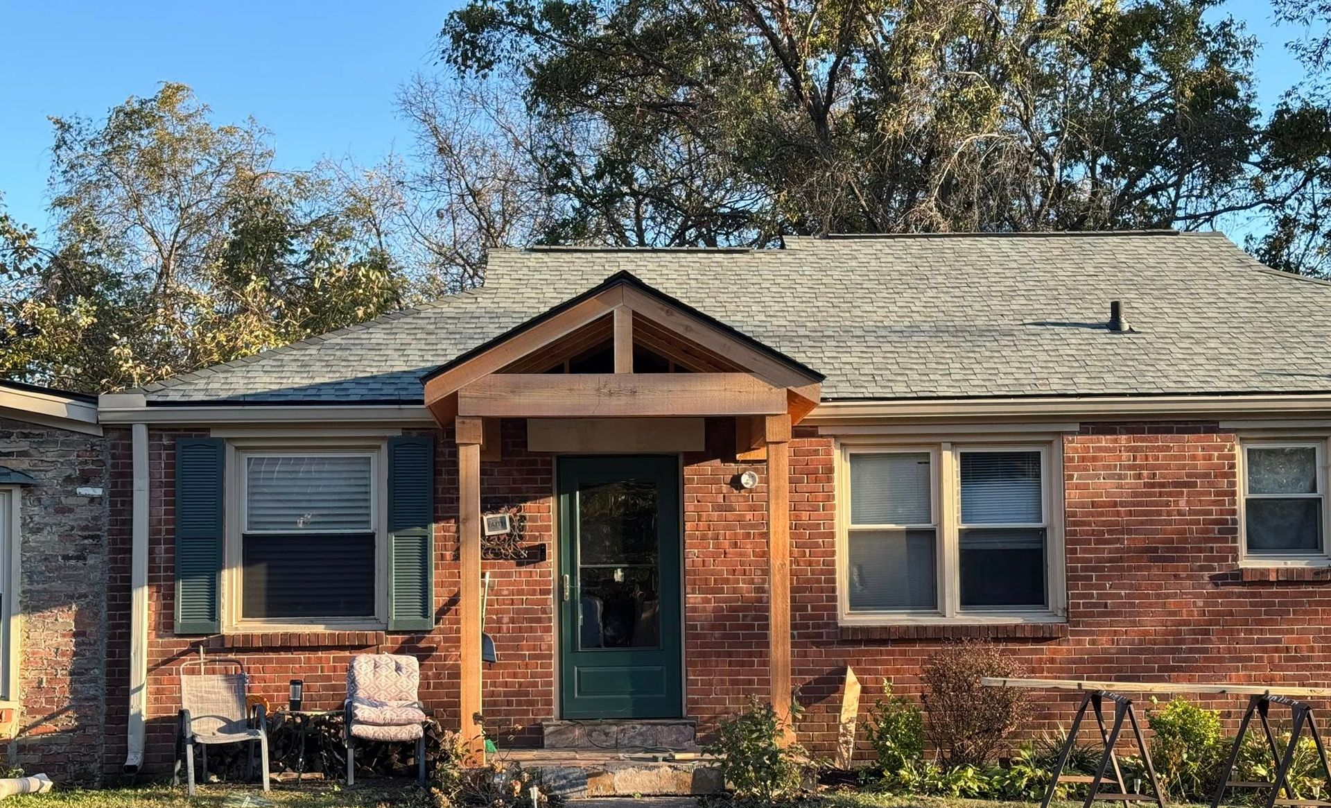 A single-story brick house with a gabled porch entrance, a green front door, and two chairs on the front patio.