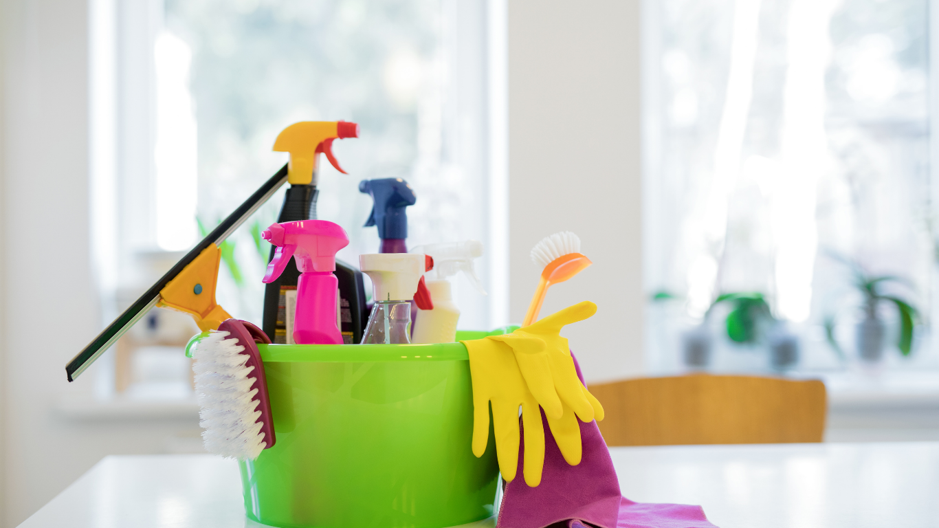 Green bucket filled with cleaning supplies: spray bottles, brush, gloves, and window squeegee sit on a table near a window.