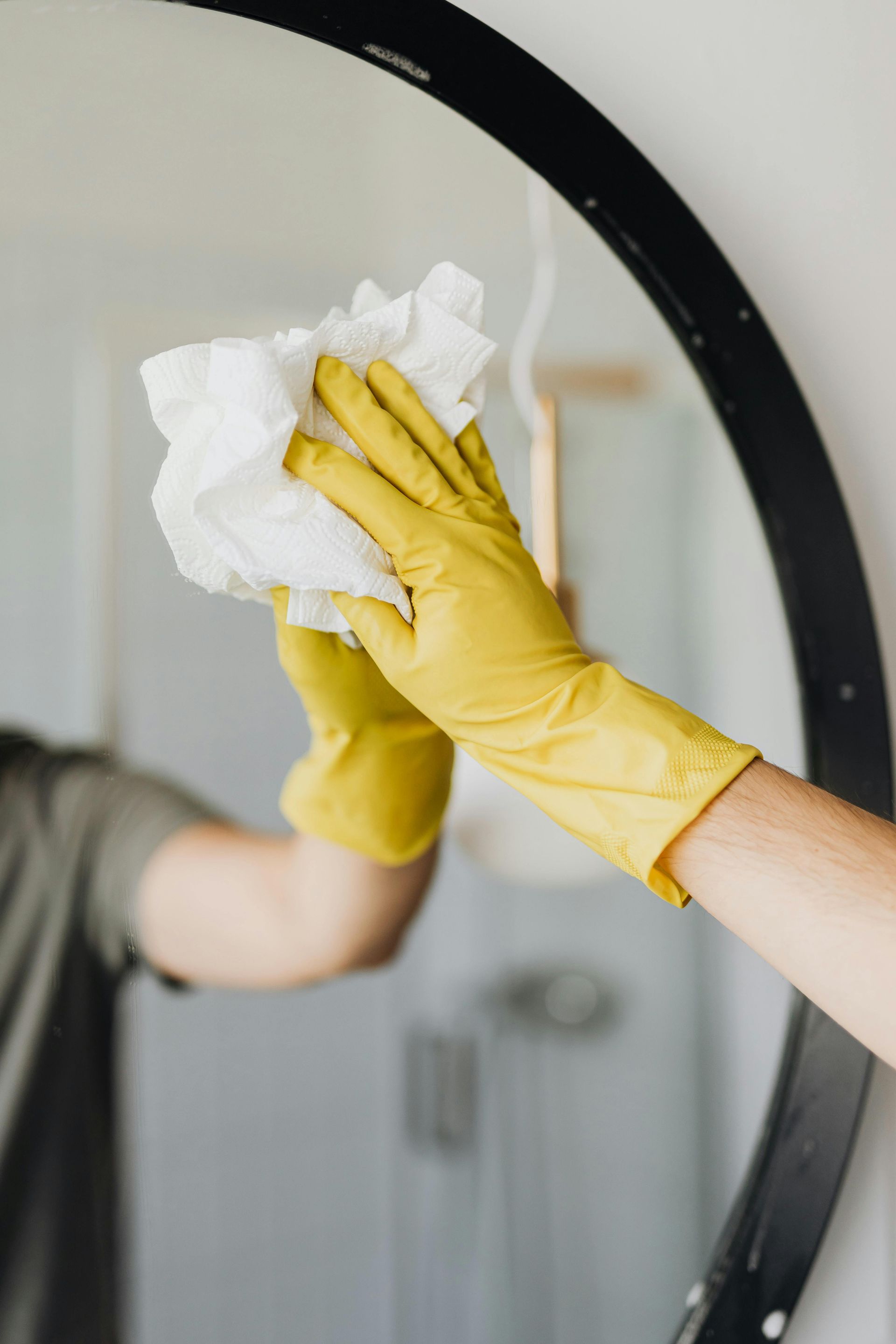Yellow-gloved hand wiping a round mirror with a white cloth. Close-up in a bathroom.