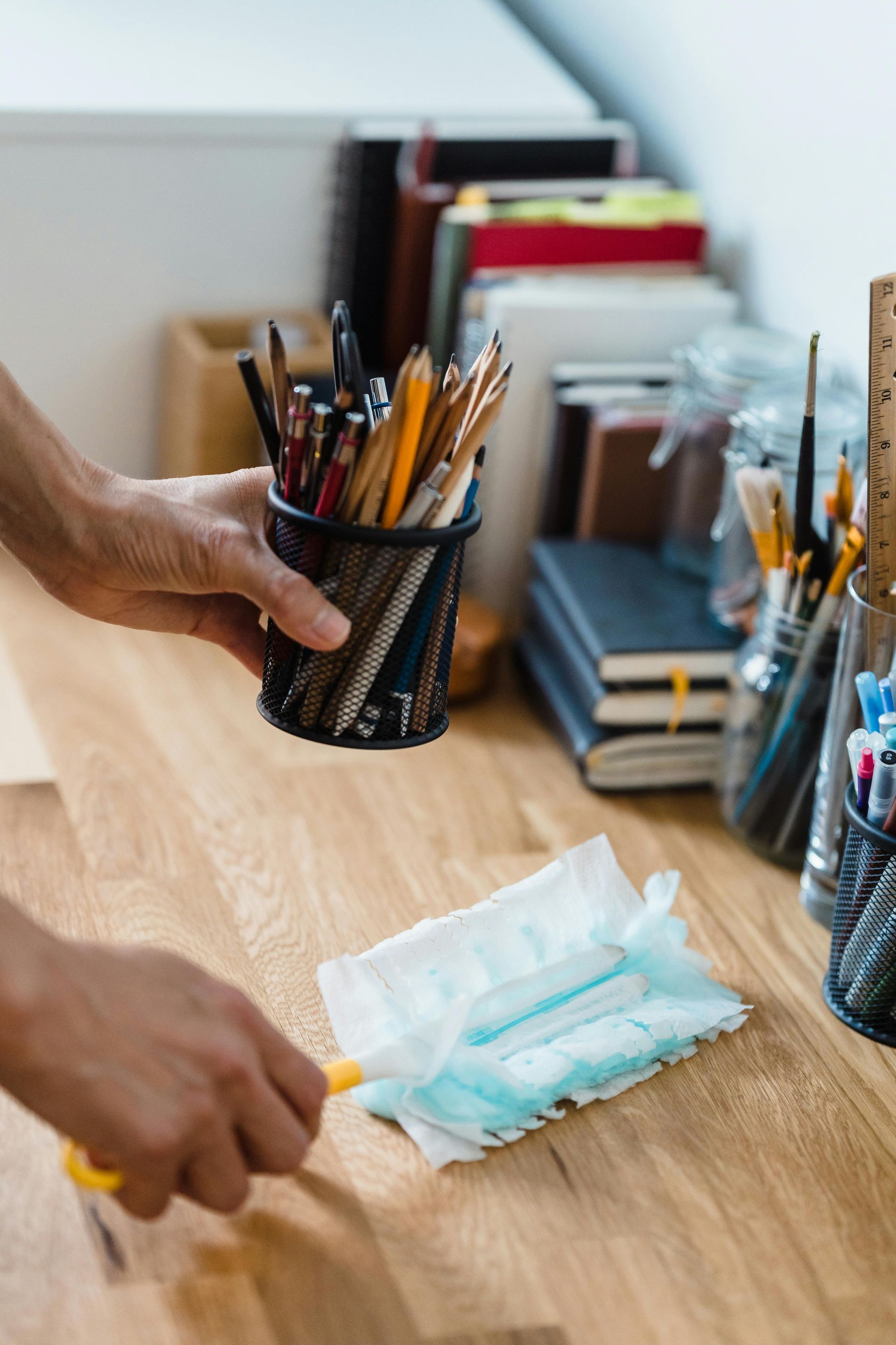 Person dusting a wooden desk with a blue duster while holding a pencil holder with pencils.