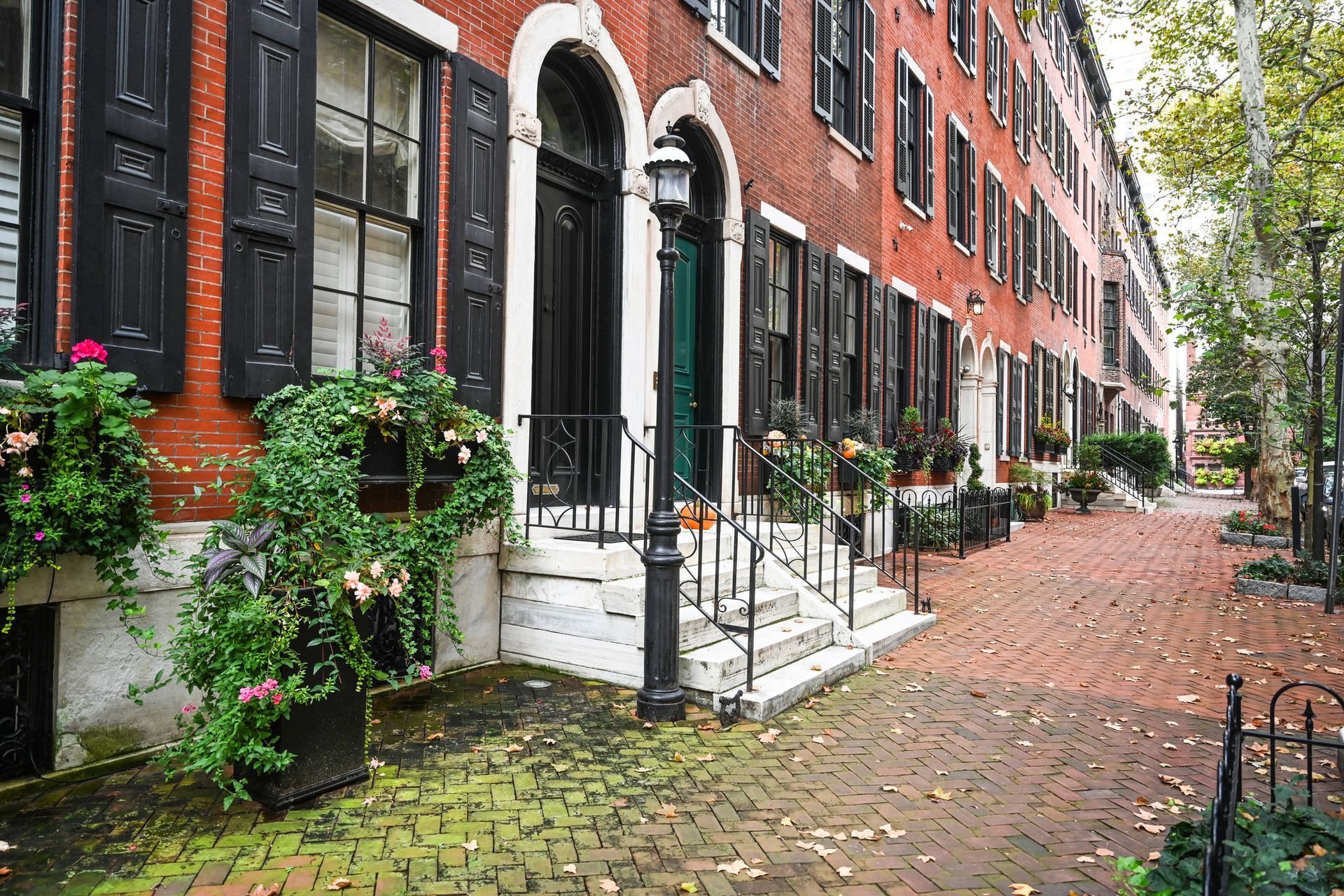 Red brick rowhouses with black shutters and flower boxes along a leafy cobblestone street Red brick rowhouses with black shutters and flower boxes along a leafy cobblestone street