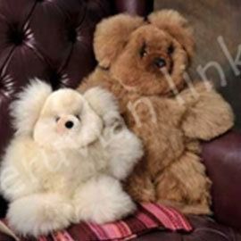 Two plush alpaca teddy bears, one cream and one brown, sit on a brown leather couch, with a striped pillow visible.
