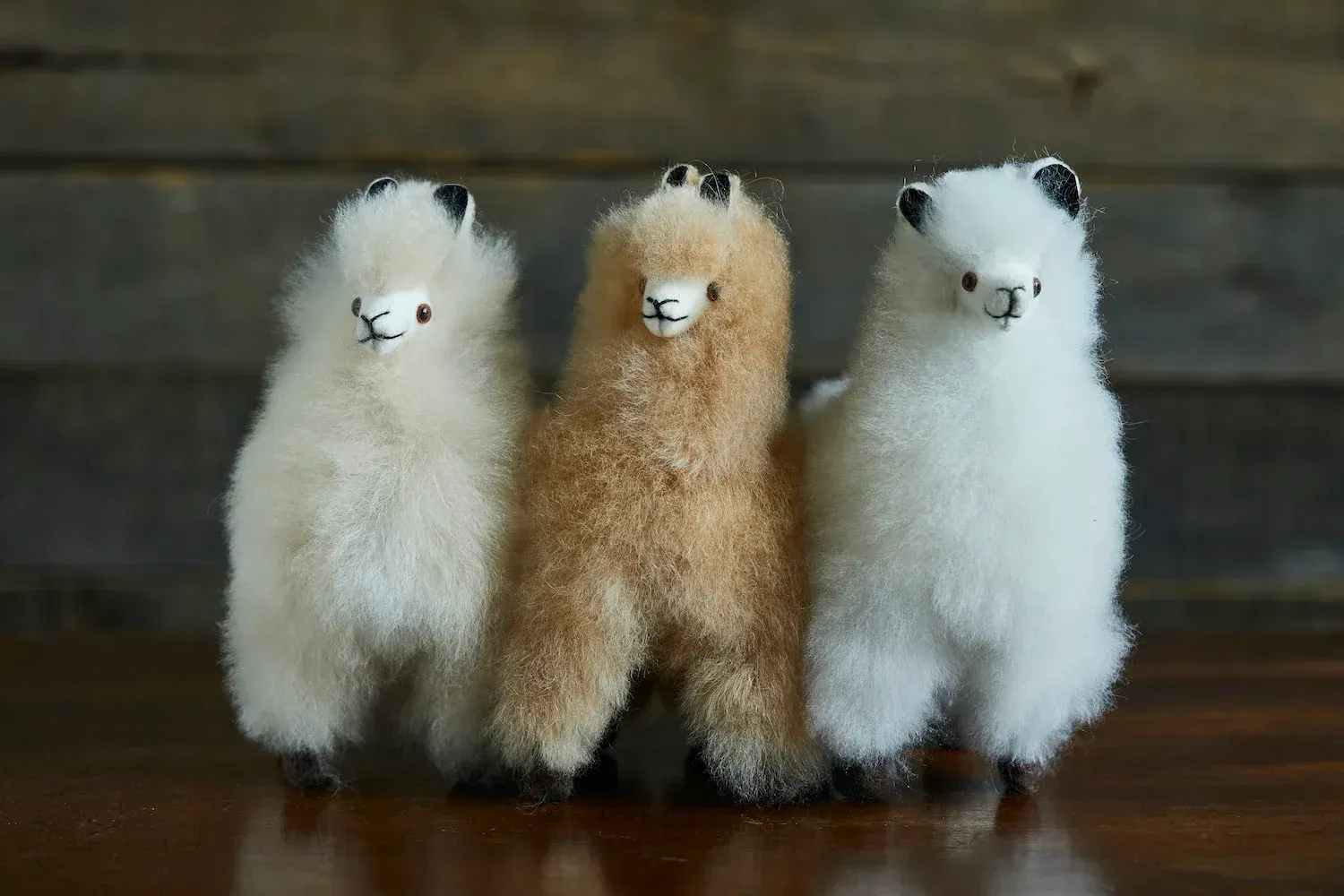 Three fuzzy alpaca figurines: off-white, tan, and white with black ears, standing against a wood background.