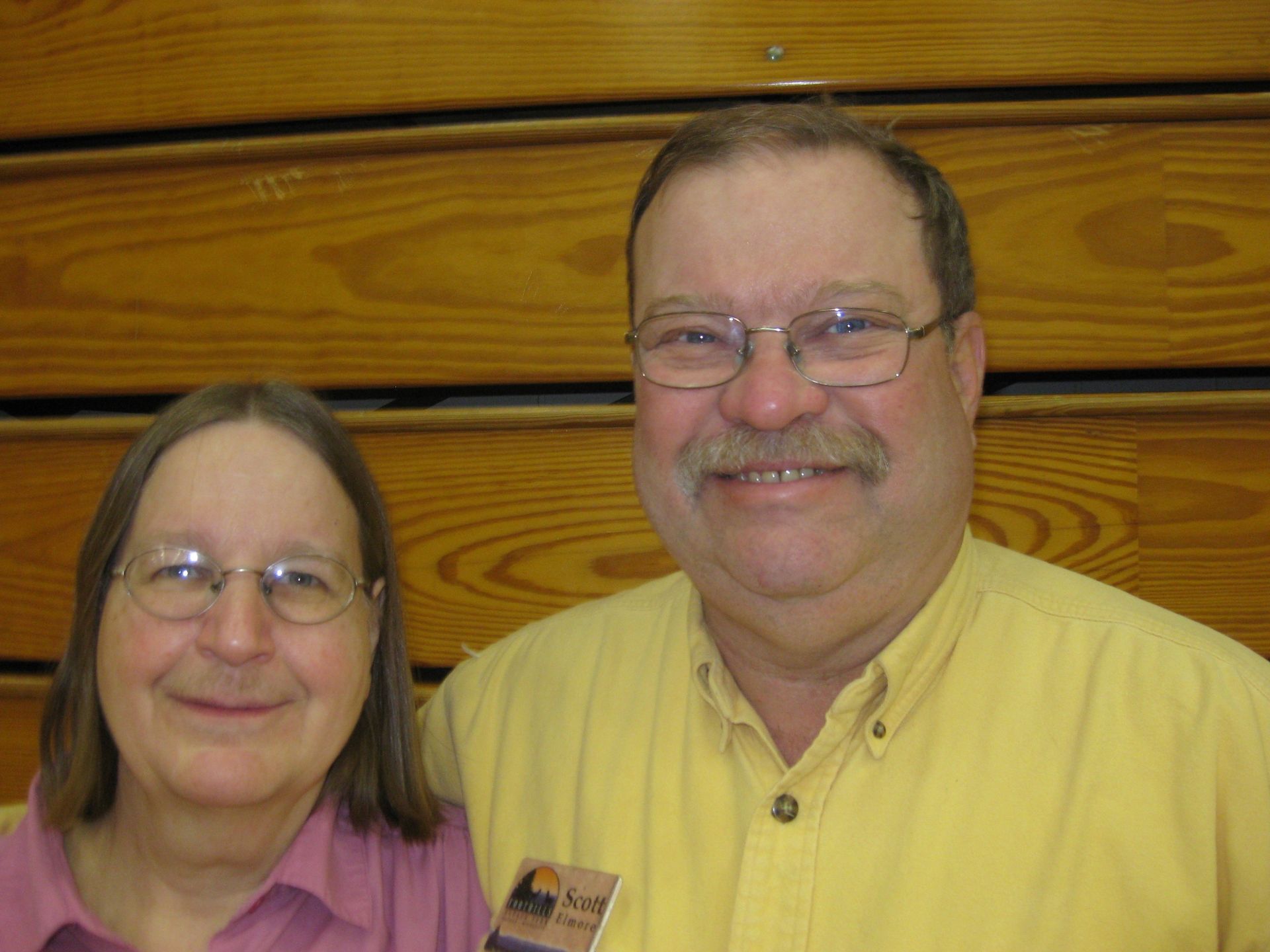 Two people, a woman and a man, smile in front of wood paneling.