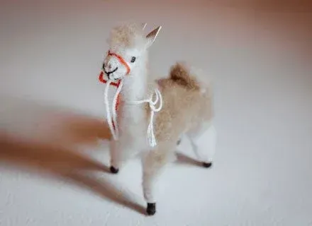 A small, fluffy llama figurine with a red bridle and white halter stands on a white surface.