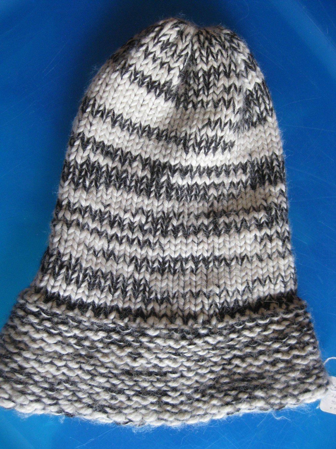 A hand-knitted hat with a brim, patterned with horizontal stripes of black, gray, and white, on a blue background.