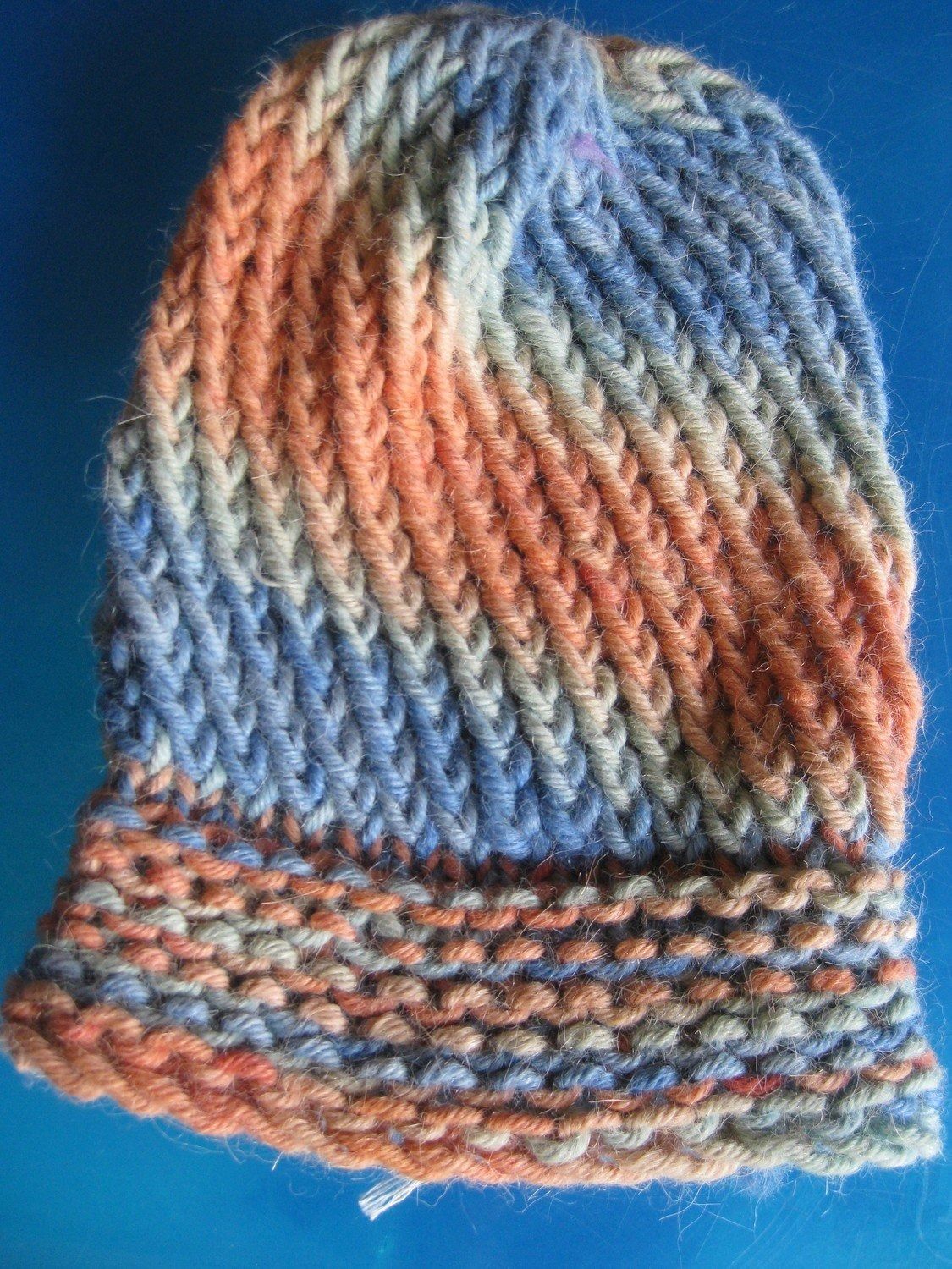 Knitted hat in a blend of orange, blue, and gray yarn, with a ribbed brim and diagonal stripes.