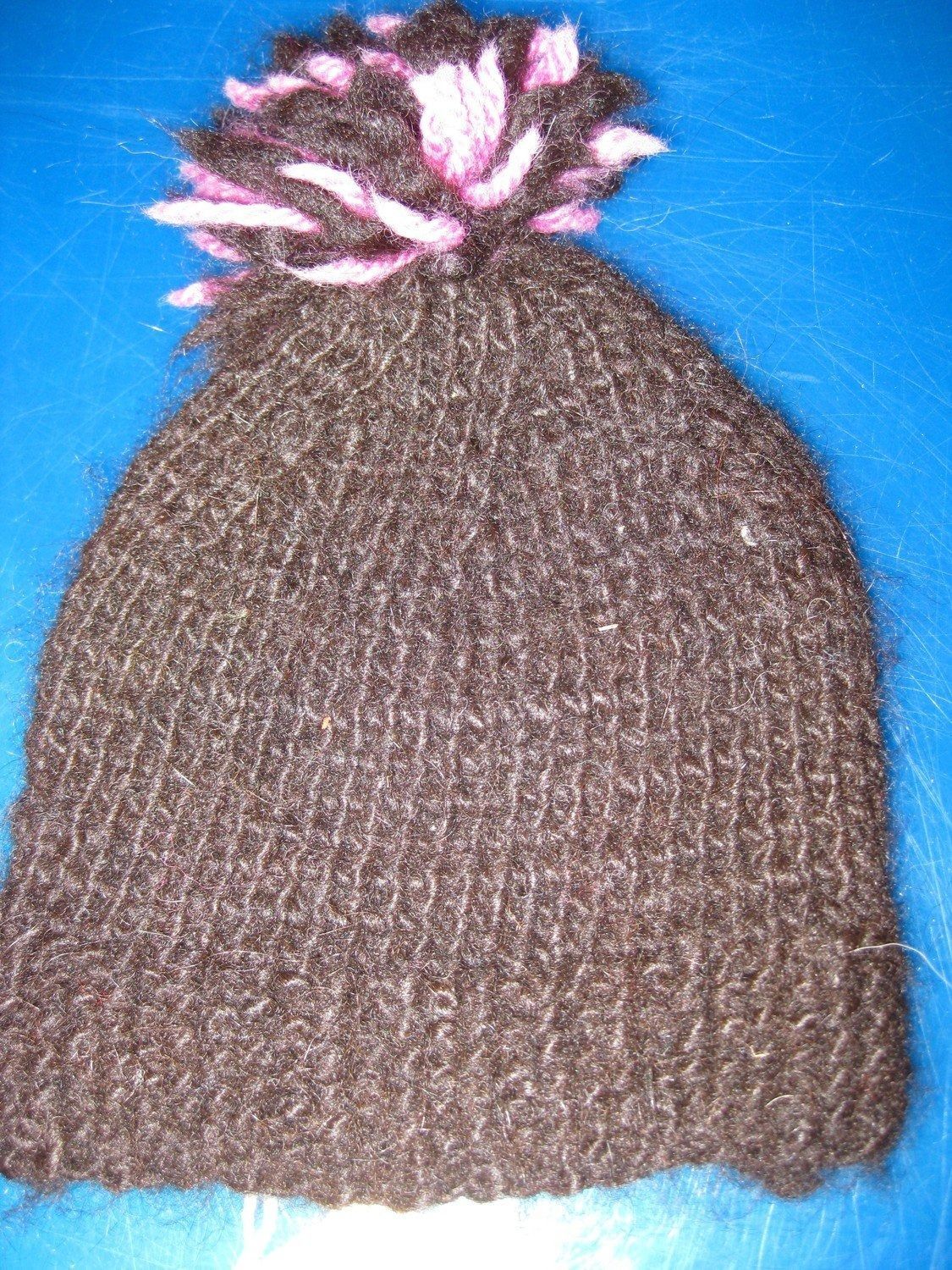 Brown knitted hat with a pink and white pom-pom on a blue surface.
