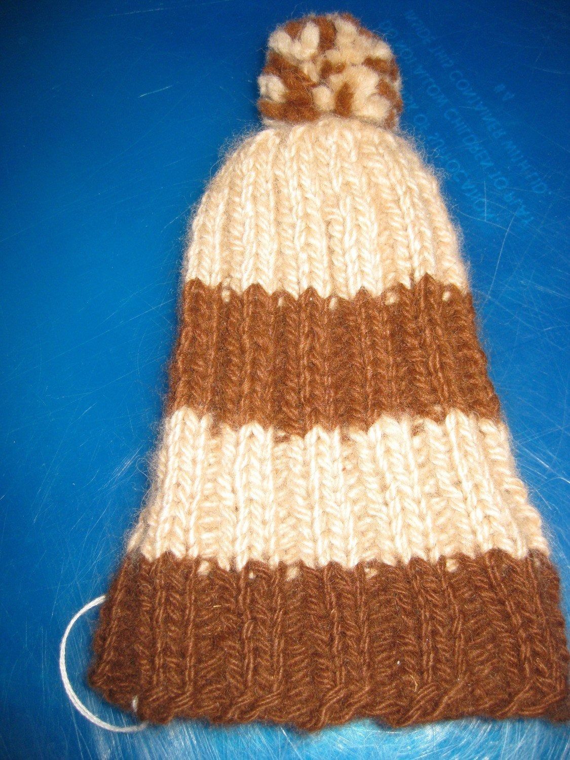 A handmade knitted hat with alternating stripes of brown and cream, topped with a brown and cream pompom.
