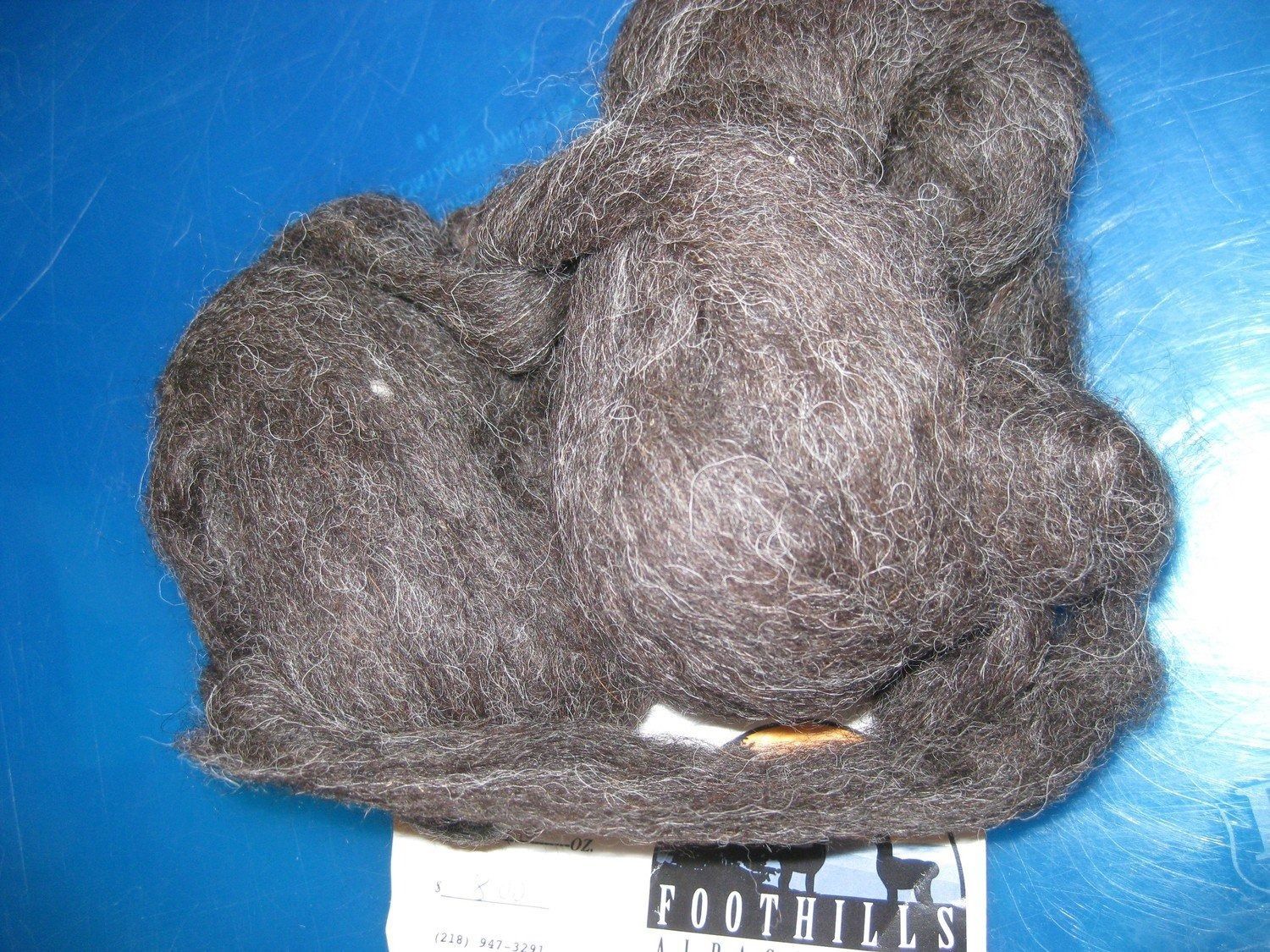 Dark brown alpaca fiber clump on a blue surface, with a label in the lower right corner.