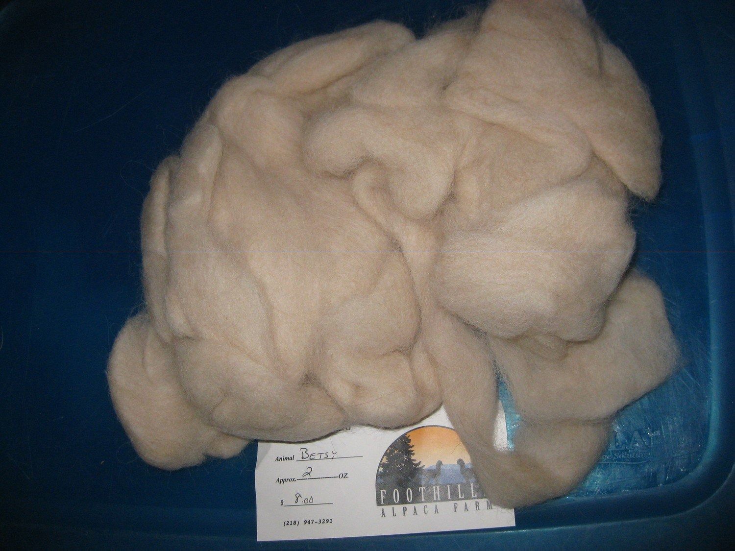A pile of light beige wool sits in a blue bin, next to a small paper with text and a logo.