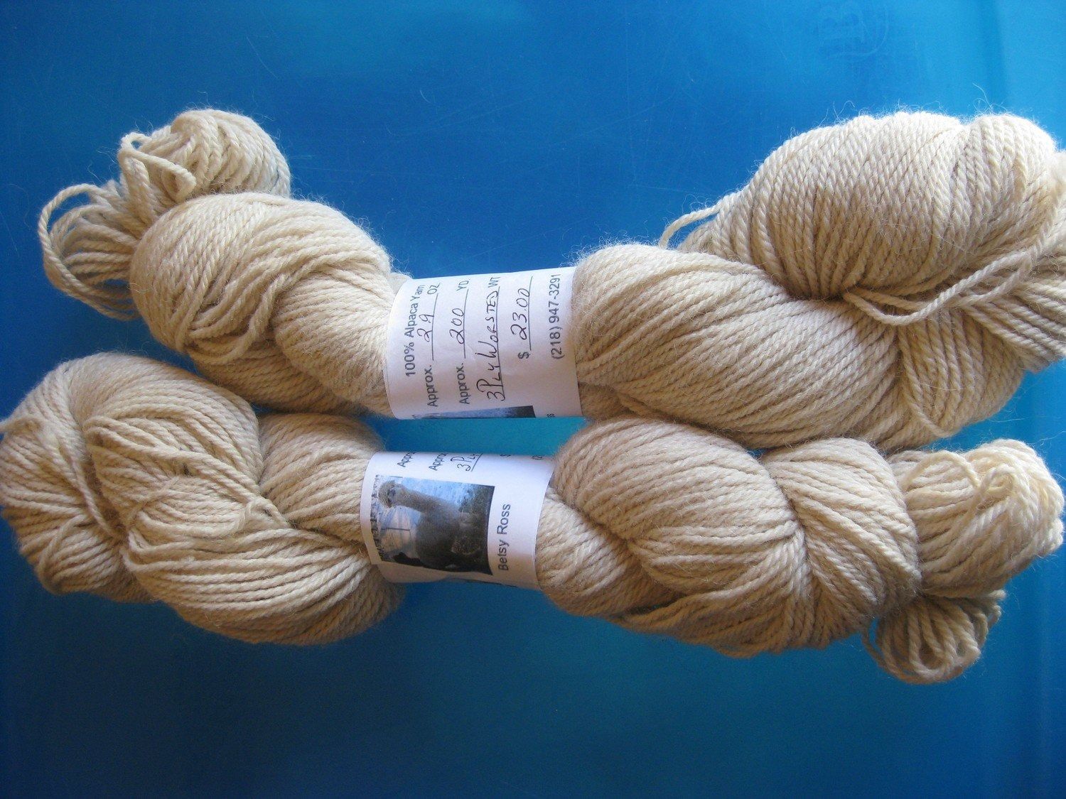 Two skeins of light beige yarn on a blue background. Each skein has a label wrapped around its center.