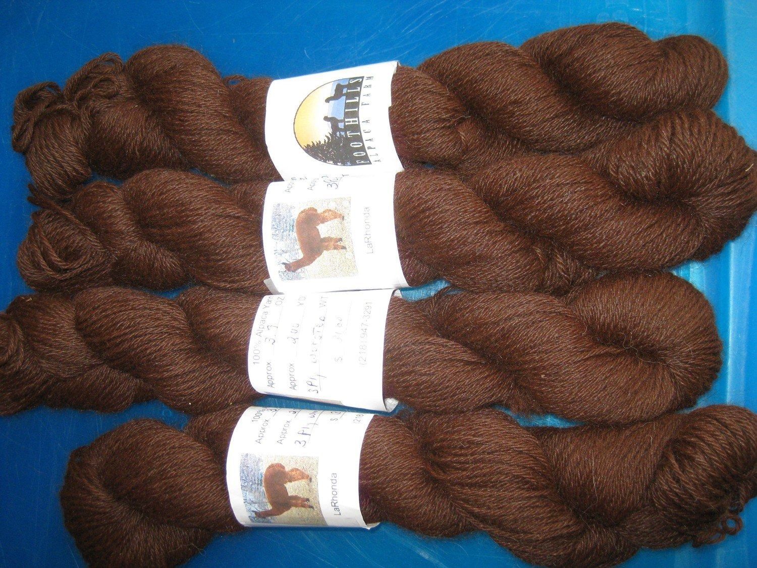 Four skeins of dark brown yarn on a blue surface, each with a white label featuring a llama silhouette and text.