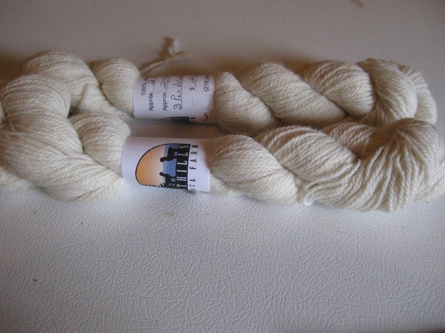 Two skeins of off-white yarn with labels wrapped around them, lying on a white surface.