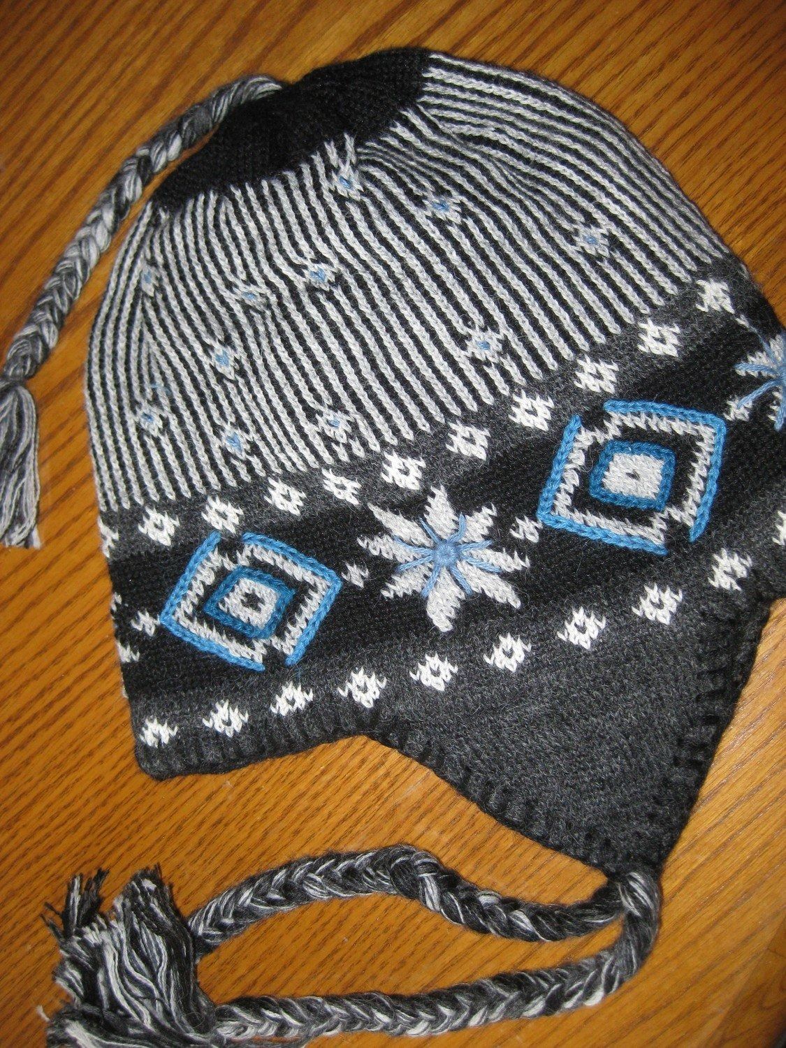 Knitted winter hat with ear flaps. Black, gray, and white design with blue accents; on a wooden surface.