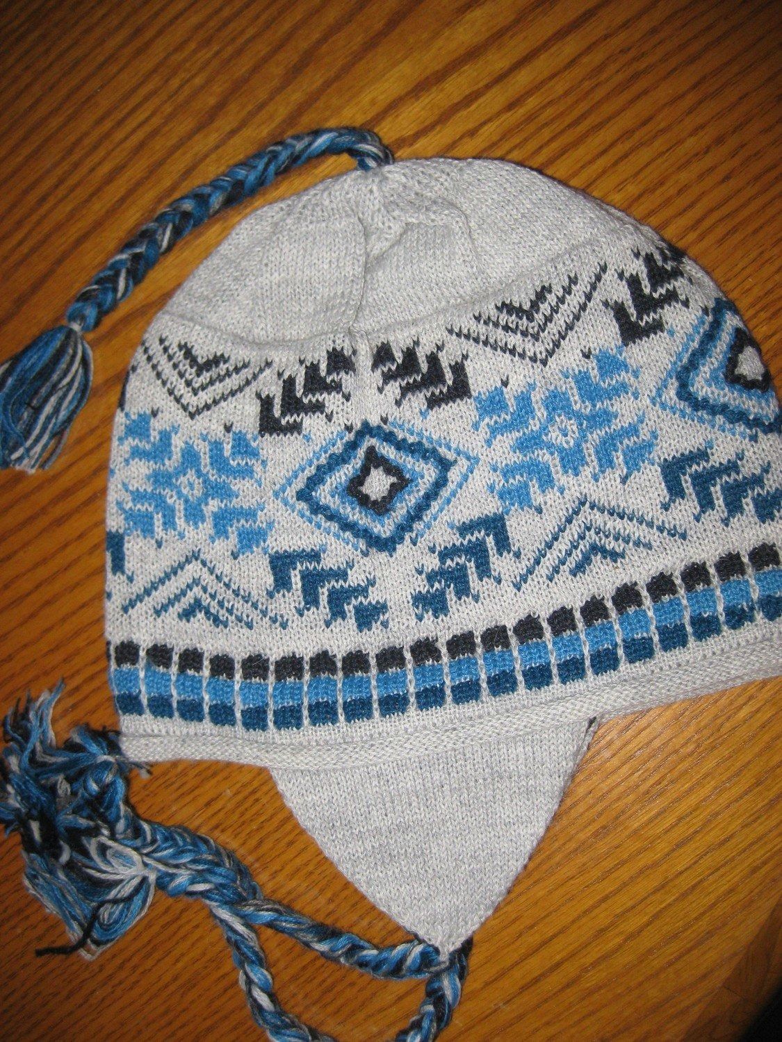 Gray knit cap with blue and black geometric patterns, ear flaps, and braided tassels.