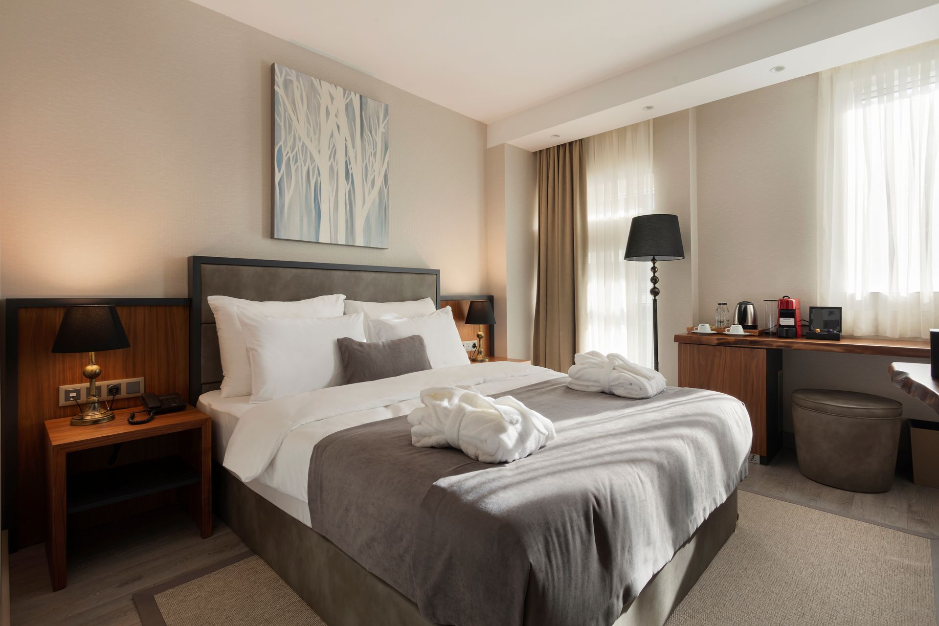 Düzgünce hazırlanmış yatak, komodinler, yer lambası ve büyük pencere perdeleri bulunan modern otel odası.