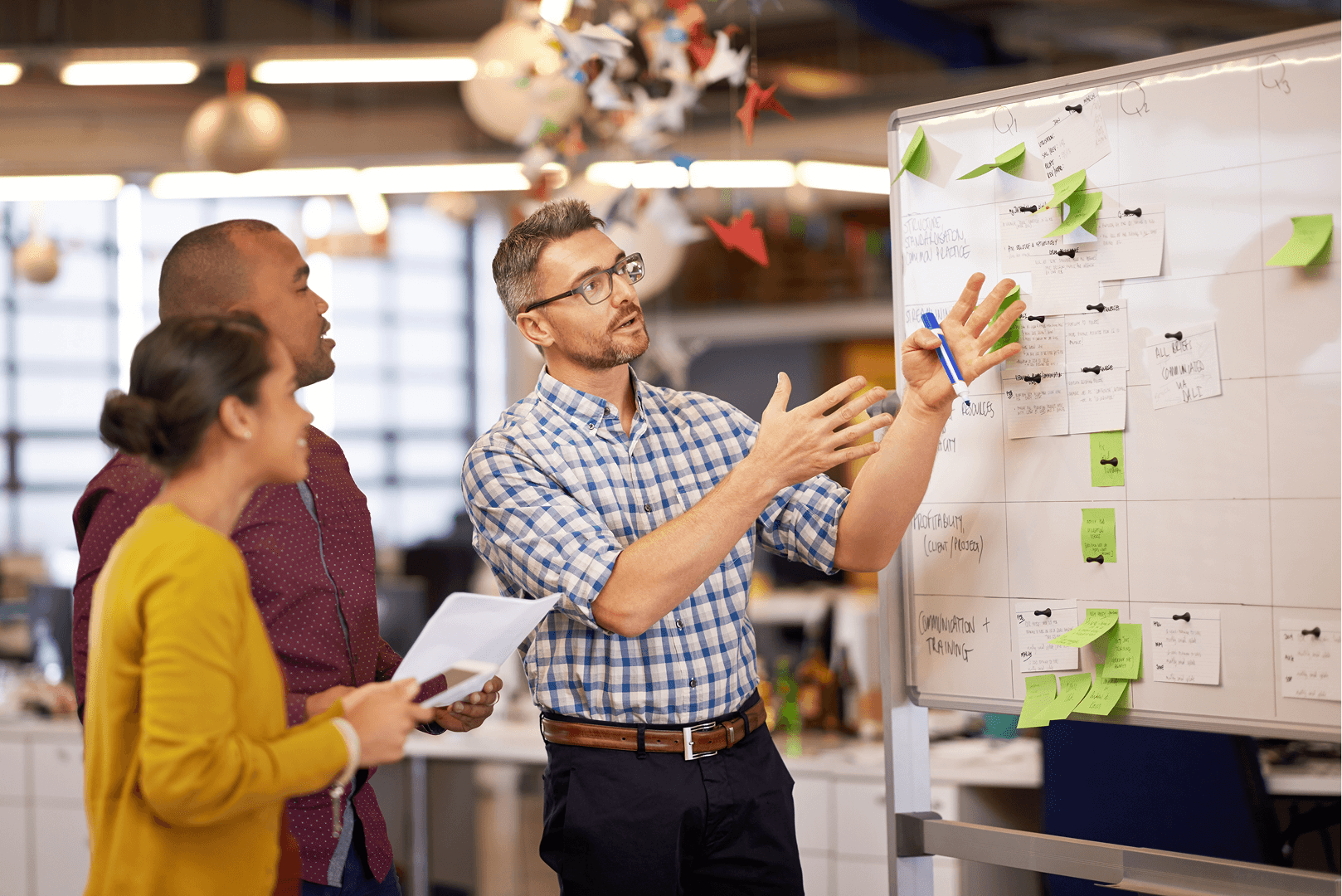 Three people collaborating at a whiteboard covered with sticky notes; man gestures with pen.