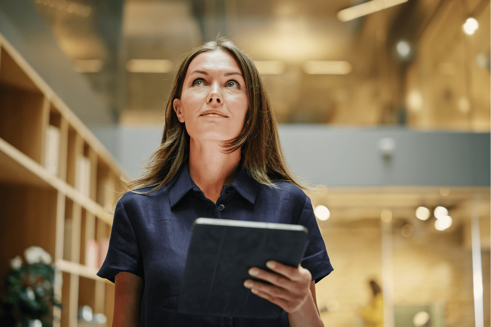 Woman in blue shirt holding tablet, looking up. Office setting, bookshelves.
