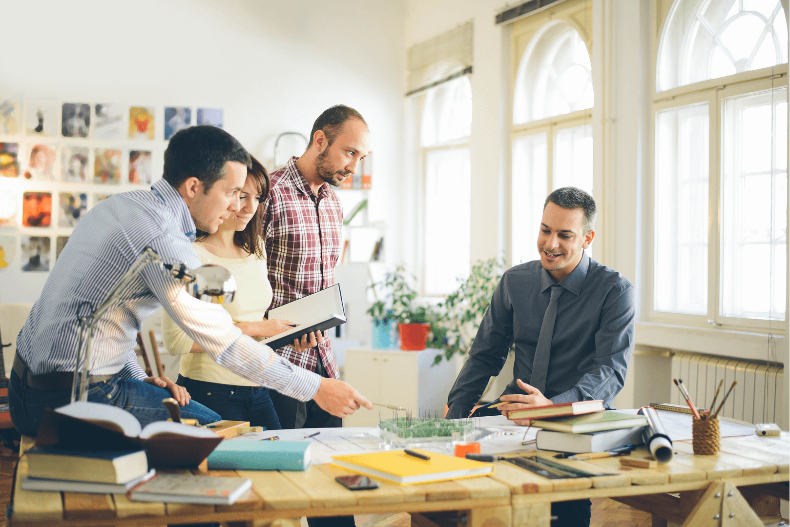 Four people at a table, discussing paperwork. Two look at papers, one points. One person sits. Bright office.