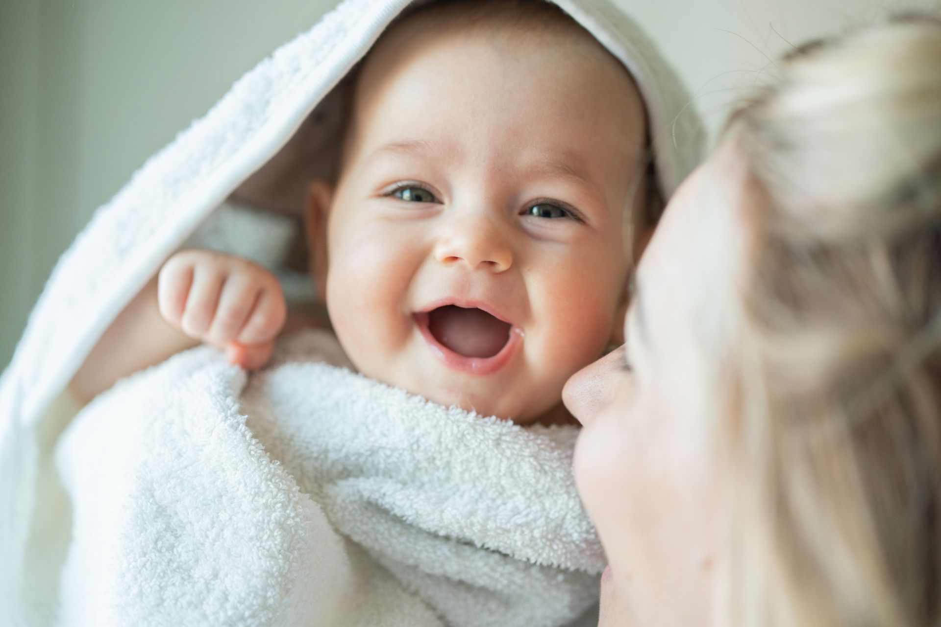 Baby wrapped in a white towel, smiling widely as a person kisses their cheek.