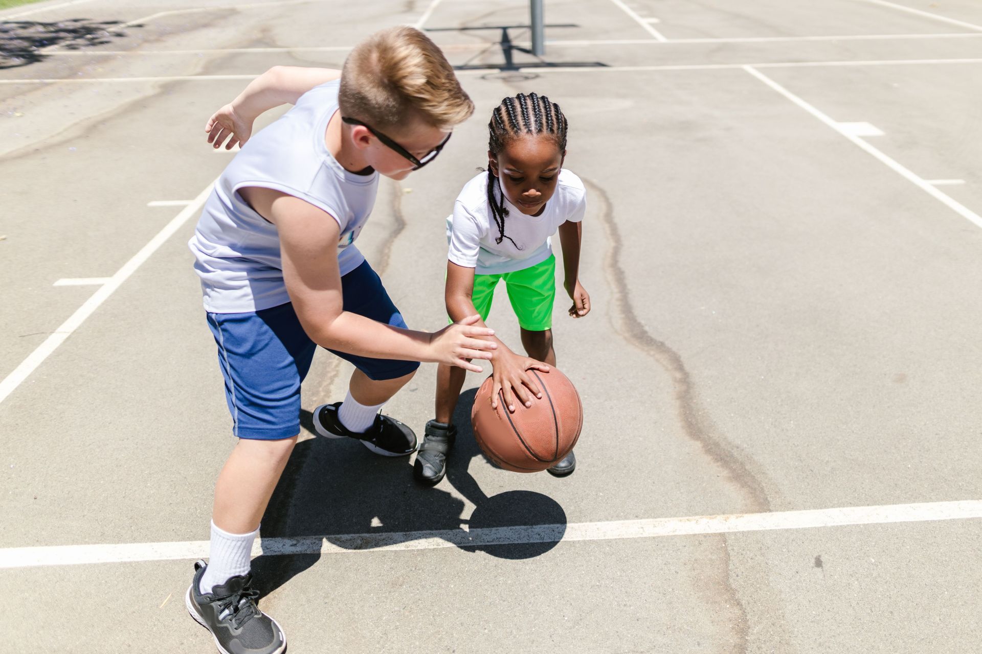 Two children dribble a basketball on an outdoor court in bright sunlight.