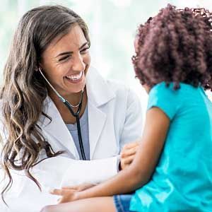 A doctor is smiling while listening to a little girl 's heartbeat with a stethoscope.