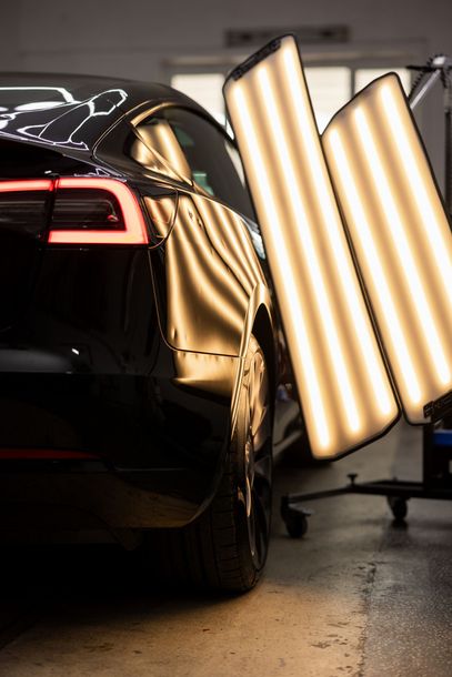 Black car in a studio beside bright vertical lights, with reflections on its glossy surface.